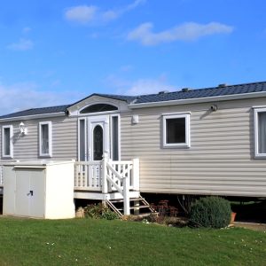 A manufactured home contains cream-colored siding, a white porch, a white entry door, windows, and a small white shed.