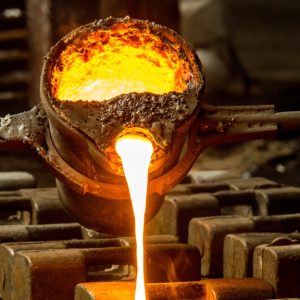 A bucket full of hot, melted metal is being poured into a cast. Several casts line up ready for the melted metal.