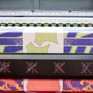 A large, black DTF printer is printing a purple, gold, and white scene of shapes onto a piece of fabric.