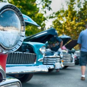 Middle-aged adults viewing classic cars at a car show. Each car has an open hood and a gleaming exterior.