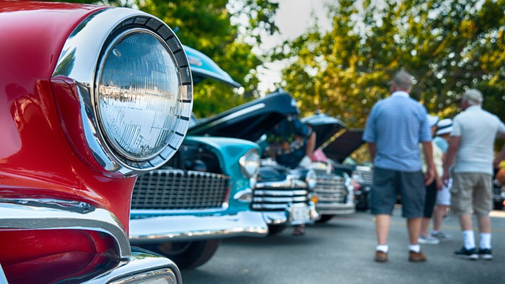 Middle-aged adults viewing classic cars at a car show. Each car has an open hood and a gleaming exterior.