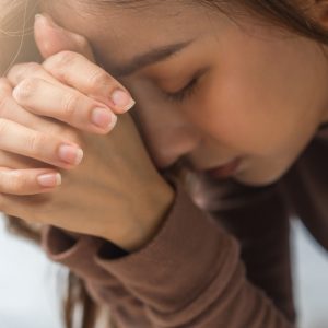 A close-up of a young woman bowing forward and leaning her head on her clasped hands in prayer.