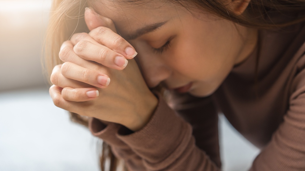 A close-up of a young woman bowing forward and leaning her head on her clasped hands in prayer.