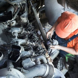 A high-angle view of a worker in overalls and an orange hat, working on the exposed engine at the front of a semi truck.