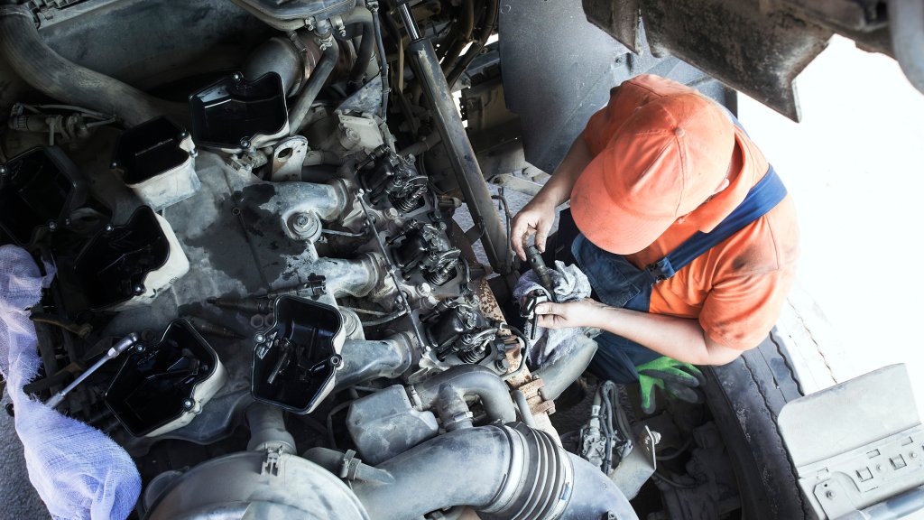 A high-angle view of a worker in overalls and an orange hat, working on the exposed engine at the front of a semi truck.