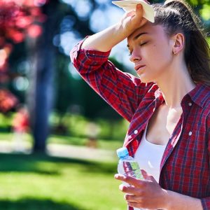 A young, tired woman standing in a beautiful park on a sunny day as she wipes her forehead sweat and holds a water.