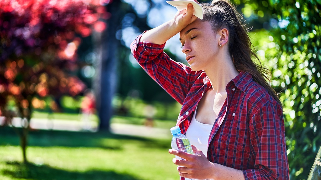 A young, tired woman standing in a beautiful park on a sunny day as she wipes her forehead sweat and holds a water.