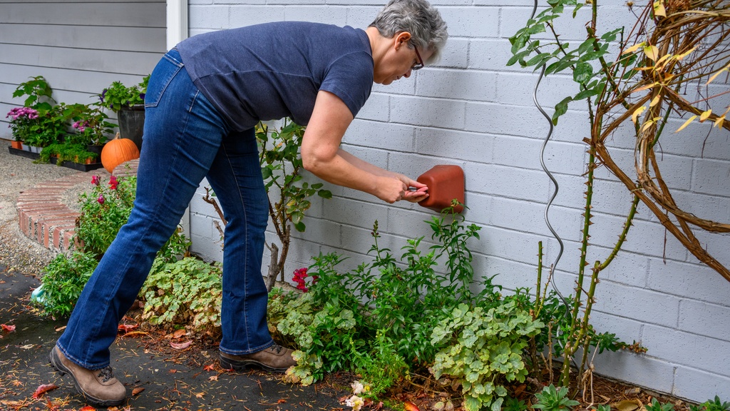 A woman with short gray hair leans forward to install a foam cover over a plumbing outlet outside a house.