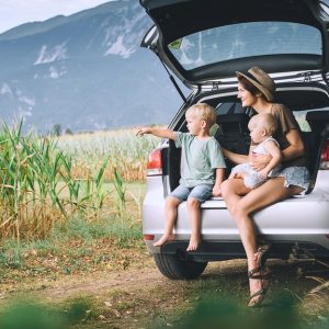 A mom and her two kids sit in the open trunk of her car, looking out at the countryside with mountains in the back.