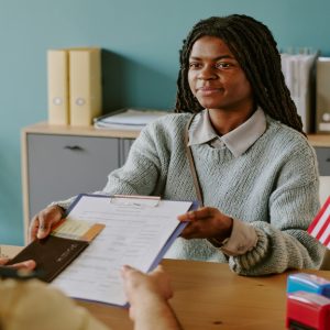 A woman hands over a clipboard of documents to a person sitting across them. On the table is the American flag.
