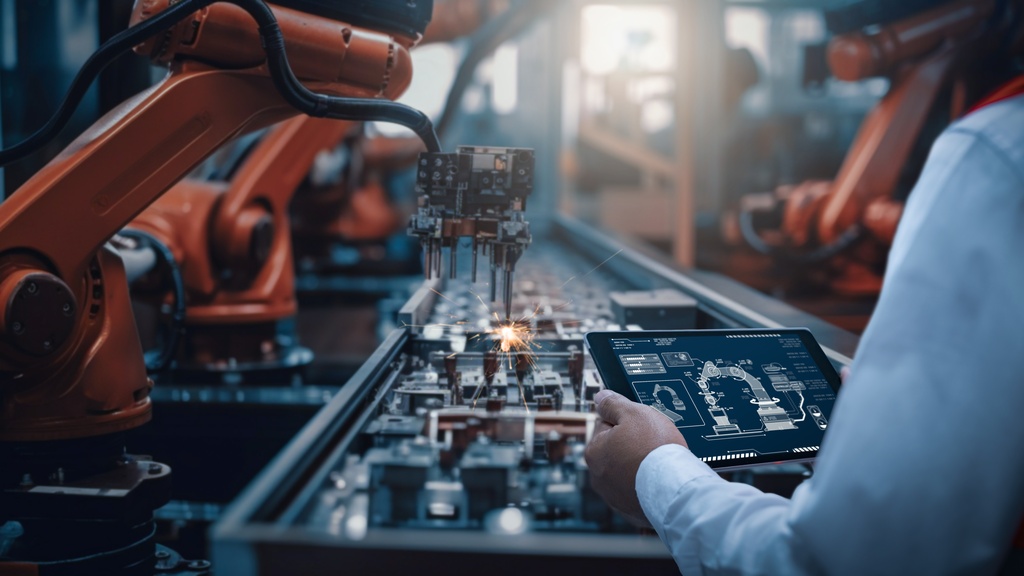 An over-the-shoulder view of an engineer examining an e-tablet and a manufacturing line with automated machines.