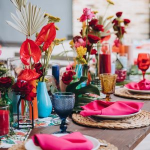 A wooden banquet table, decorated with tropical-style decor and flowers. Colorful napkins, candles, and flowers are on the table.