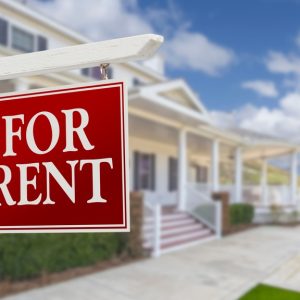 A red "For rent" sign in the front of a rental property. The sidewalk is groomed with a white porch and cement steps.