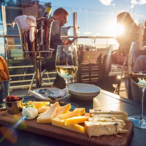 People sit at a table with a wooden board with various cheeses and toasted bread, along with glasses filled with white wine.