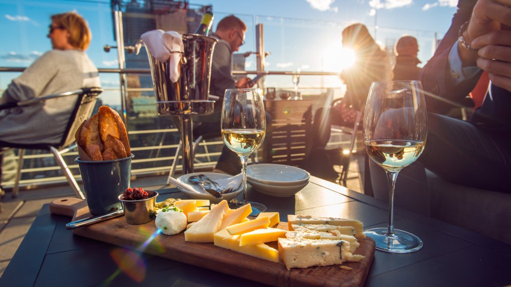 People sit at a table with a wooden board with various cheeses and toasted bread, along with glasses filled with white wine.