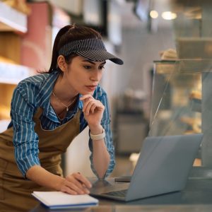 A woman bends over a bakery counter, looking at a laptop with one hand under her chin and the other resting on a small notepad.