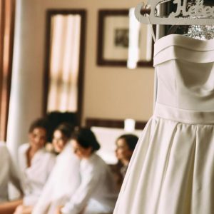 A wedding gown hanging on a customized name hanger on the mirror. The bridal party sits behind it in white robes.