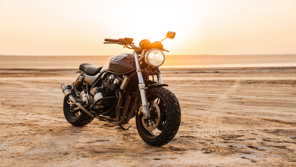 A motorcycle parked on a sandy surface, with the sun setting behind it. There is one circular headlight on the front.
