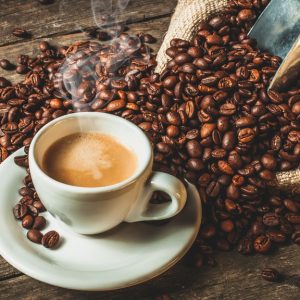 A cup of coffee with cream mixed in and steam coming from the top sits on a wooden table next to spilled coffee beans.