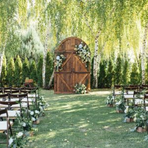 An outdoor wedding ceremony space beneath a willow tree. There is a large arched wooden door decorated with a lot of flowers.
