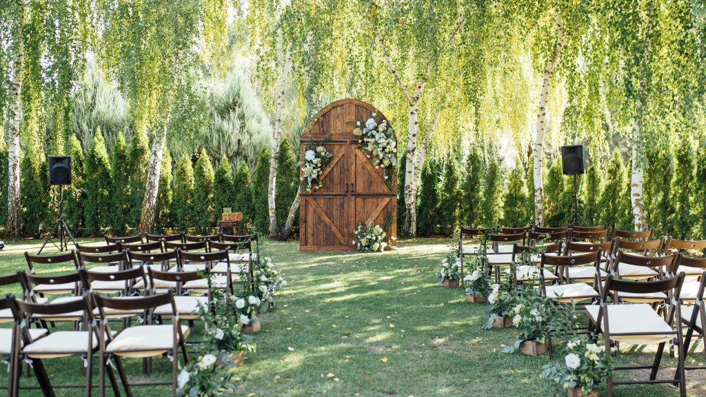 An outdoor wedding ceremony space beneath a willow tree. There is a large arched wooden door decorated with a lot of flowers.
