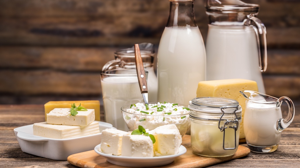 A collection of dairy products sit on a wooden table. The products range from milk, cheddar cheese, and cottage cheese.