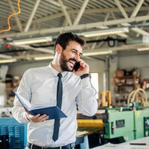 A smiling man stands in a print production room talking on a cell phone and holding an open book.