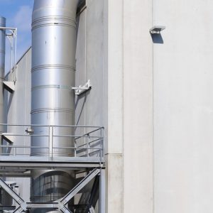 Large metal pipes come out of the side of a cream-colored manufacturing building. The blue sky appears in the background.