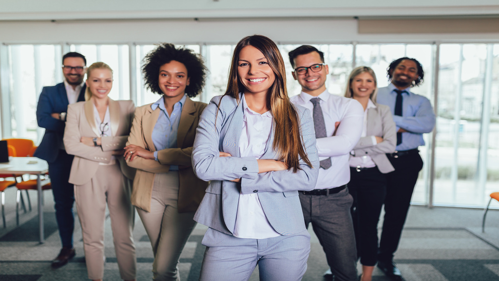 A group of people dressed in formal clothes and smiling, standing in an pointed formation inside an office environment.