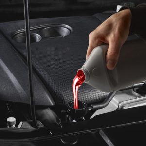 An individual in a black sweatshirt pours bright red liquid from a gray bottle into a car engine's black funnel.