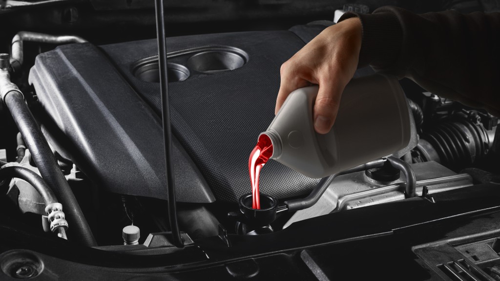 An individual in a black sweatshirt pours bright red liquid from a gray bottle into a car engine's black funnel.