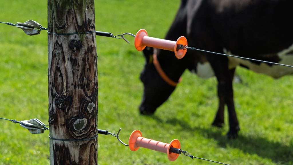 A wooden post with orange electric fence handles and wires in front of a grazing black-and-white cow in a green pasture.