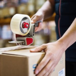 A person seals a brown cardboard box with a red and grey tape dispenser in a warehouse filled with stacked boxes.