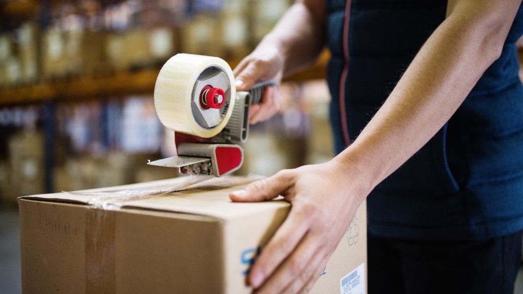 A person seals a brown cardboard box with a red and grey tape dispenser in a warehouse filled with stacked boxes.