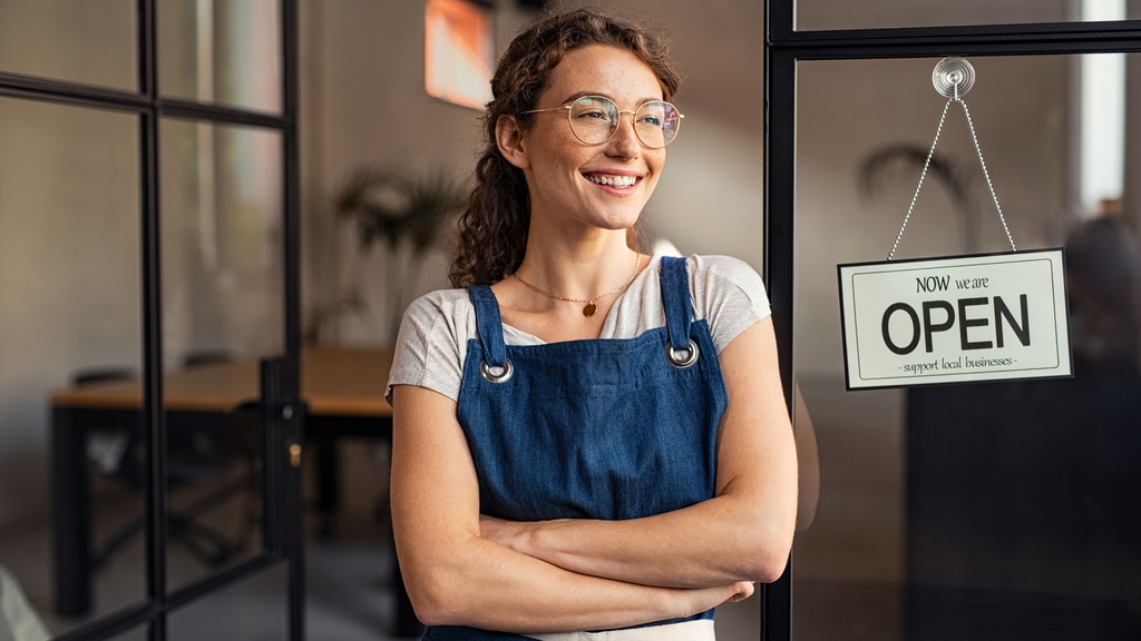A woman wearing glasses and a blue apron smiles in front of a sign that says "Now we are OPEN" and "Support local businesses."s