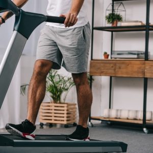 A man in a white T-shirt and gray shorts grips the handlebars of a treadmill while walking on an incline.