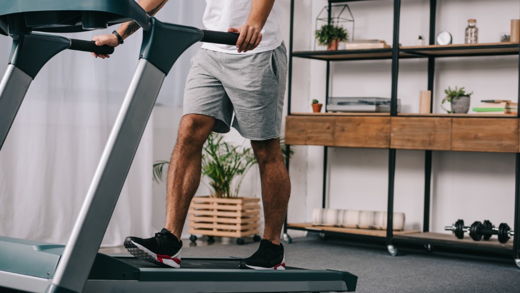 A man in a white T-shirt and gray shorts grips the handlebars of a treadmill while walking on an incline.