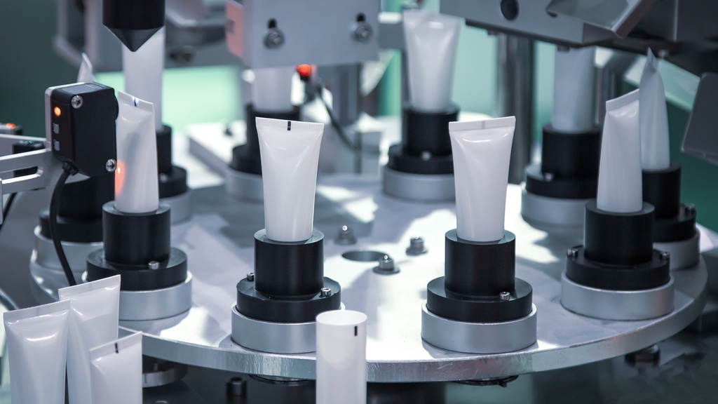 An automated machine fills and seals cosmetic plastic tubes on a rotating platform in a high-tech manufacturing facility.
