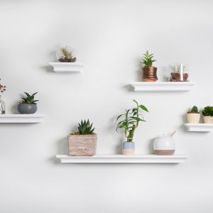 A white wall in a room with five white floating shelves staggered displaying home decor pieces and trinkets.
