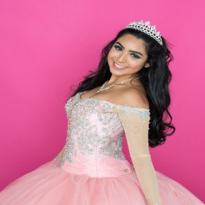 A smiling young woman in a pink embellished ball gown and tiara, posing against a bright pink background.