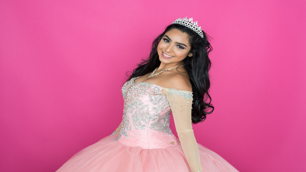 A smiling young woman in a pink embellished ball gown and tiara, posing against a bright pink background.
