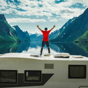 A man standing on top of an RV with his hands extended in the air. There is a lake and a mountain in front of him.