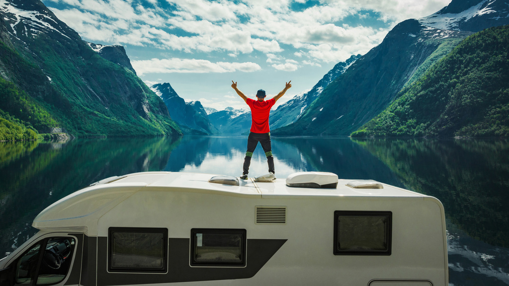 A man standing on top of an RV with his hands extended in the air. There is a lake and a mountain in front of him.
