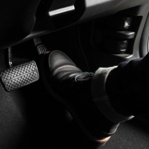 A close-up of the footwell of a driver-side cockpit of a car with a black boot pressing down on the accelerator pedal.