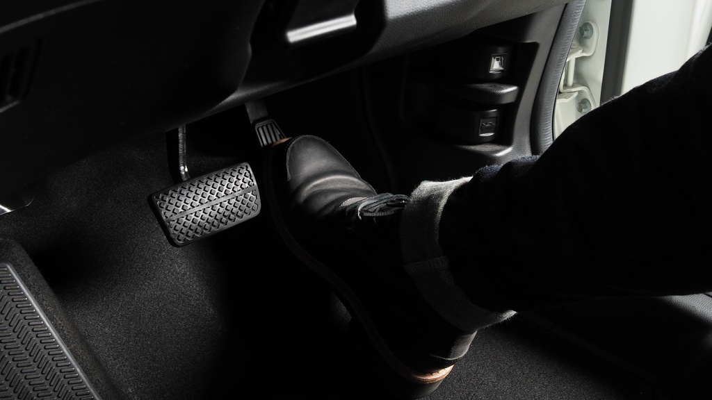 A close-up of the footwell of a driver-side cockpit of a car with a black boot pressing down on the accelerator pedal.
