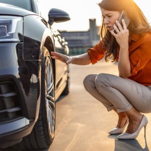 A well-dressed woman knelt next to her car, looking at her tire while on the phone. She has her hand on the tire.