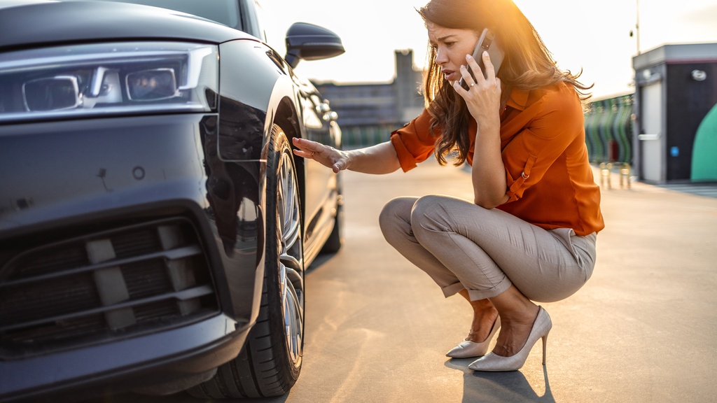 A well-dressed woman knelt next to her car, looking at her tire while on the phone. She has her hand on the tire.