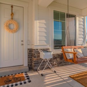 The front porch of a home features a swing, a basket, a wreath on the door, and a welcome mat featuring a motorcycle.