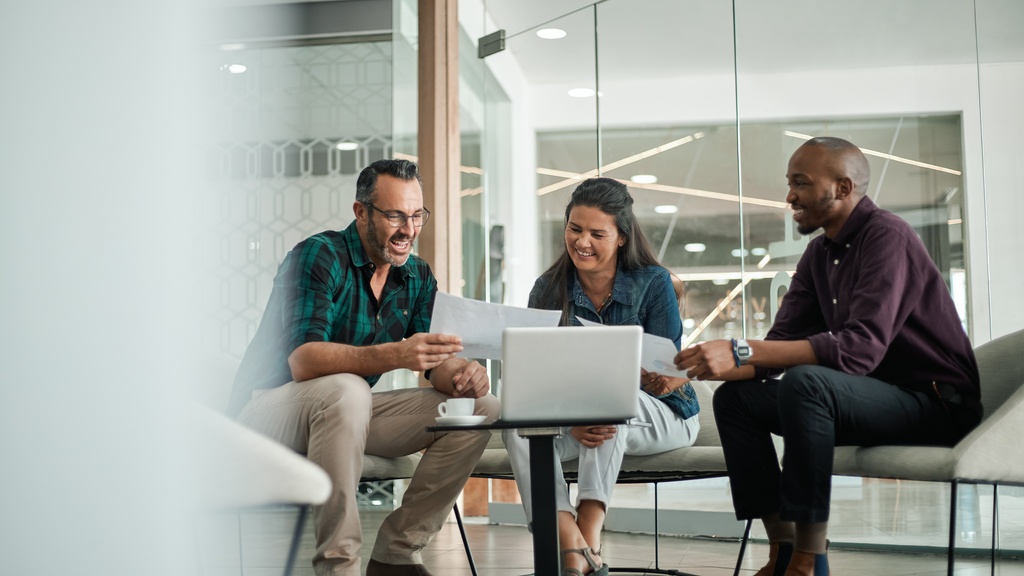 A business meeting between three people in a modern office space. They are looking at papers and a laptop together.