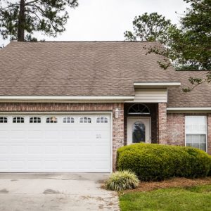 The front of a small brick house with a wide slanting roof. The driveway to the home is covered in dirt.